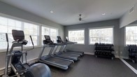 This single-floor fitness center is thoughtfully designed to provide a comprehensive exercise environment. The space is anchored by a row of modern treadmills equipped with digital display consoles, ideal for cardio training at any intensity level. Additional cardio equipment, including elliptical-style machines, lines the perimeter, offering variety for full-body workouts. A multi-tiered dumbbell rack stocked with a wide range of free weights provides ample options for strength training.

The room is finished with durable, dark fitness flooring that offers both comfort and stability during exercise. Walls are painted in a cool, neutral gray tone that lends the space a clean and focused atmosphere. Recessed lighting is complemented by a ceiling fan, which aids in air circulation and comfort during intense workouts.

Natural light is a standout feature of this fitness center, with multiple large windows fitted with white horizontal blinds lining the walls on two sides. These windows allow generous daylight to fill the room while maintaining privacy, creating an open and energizing environment. The overall layout is practical and uncluttered, maximizing usable floor space and ensuring easy movement between equipment stations.