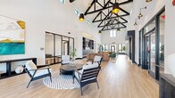 Located at 1830 Autumn Dr in Hoffman Estates, IL, this single-floor communal space offers a generously sized, open-plan interior defined by striking architectural character. The most commanding feature is the soaring vaulted ceiling with exposed black steel truss work, which lends an industrial-modern aesthetic while drawing the eye upward and amplifying the sense of volume throughout the space.

Light-toned hardwood flooring extends across the entire floor, complementing the crisp white walls and creating a bright, welcoming atmosphere. Large black-framed windows and glass entry doors flood the interior with abundant natural light and provide views of the surrounding exterior. Clerestory windows positioned high along the roofline further enhance the luminosity of the space.

The interior is organized into distinct lounge and seating areas, each furnished with contemporary chairs, round coffee tables, and patterned area rugs that define individual zones without enclosing them. A reception or leasing desk anchors one section of the floor, accompanied by built-in navy cabinetry with display shelving. A glass-walled room, visible from the main area, appears to function as a fitness or private meeting space. Pendant lighting with gold-toned interiors hangs from the trusses, adding warmth and a refined finish to the overall modern, inviting ambiance.