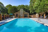 Located at 777 Valleybrook Crossing in Decatur, Georgia, this outdoor amenity area centers around a generously sized swimming pool with crystal-clear blue water. The pool is framed by a broad, stone-paved deck that provides ample space for relaxation and recreation. Surrounding the pool are multiple lounge chairs and chaise seating, offering comfortable spots for sunbathing or unwinding poolside.

A dedicated seating area on the patio features a round table with chairs shaded by a vibrant blue umbrella, ideal for outdoor dining or casual gatherings. The pool area is enclosed by a classic wrought iron fence supported by brick columns, lending a refined and secure feel to the space.

A neighboring pool house or clubhouse structure with a pitched roof and chimney is visible at the perimeter, adding architectural character to the setting. The entire area is beautifully framed by mature, leafy trees that provide natural shade and a sense of privacy, creating a tranquil retreat-like atmosphere. The lush greenery backdrop contrasts warmly with the stone hardscape and the vivid pool water, resulting in an inviting and well-appointed outdoor space suited for warm-weather enjoyment.