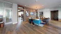 This thoughtfully designed single-floor space blends industrial character with contemporary comfort across several distinct zones. Exposed brick accent walls serve as a defining architectural feature throughout, creating a warm, textural contrast against clean white walls and smooth hardwood flooring that extends throughout the entire space.

The main lounge area features an open, airy layout anchored by bold, curated furnishings, including a vibrant teal sofa, leather accent chairs, and eclectic artwork. A bar-height communal table sits near floor-to-ceiling windows and a retractable glass wall, flooding the space with natural light and providing views of the surrounding outdoor area.

A dedicated dining nook offers a rustic wood communal table paired with navy velvet upholstered chairs, set against a striking geometric patterned wallcovering with an industrial-style pendant light. A quieter lounge alcove features neutral-toned modular seating arranged around a glass coffee table, ideal for more intimate gatherings.

The kitchenette area is equipped with navy cabinetry, a stainless steel dishwasher, a white marble-style island, and globe pendant lighting. Glass-partitioned conference or flex rooms are visible throughout, offering semi-private spaces for work or meetings, rounding out the versatile functionality of this well-appointed communal space.