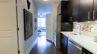 This single-floor space is thoughtfully designed to maximize functionality and style. A long hallway serves as the central corridor, featuring rich dark hardwood flooring that extends throughout the unit, creating a cohesive and elegant aesthetic. The left wall of the hallway is decorated with framed wall art, including a notable map print, alongside decorative kitchen-themed accents.

The galley-style kitchen lines the right side of the corridor, outfitted with dark espresso cabinetry that reaches toward the ceiling, providing generous storage. Light stone countertops offer a striking contrast against the dark cabinetry, and stainless steel appliances, including a French door refrigerator and sink, complete the modern kitchen setup.

At the end of the hallway, a louvered door opens into a bright living area that benefits from abundant natural light streaming through large windows. The living space features a ceiling fan with a light fixture and offers views of the surrounding cityscape. The overall ambiance of the space balances contemporary design elements with warm, inviting tones, creating a comfortable and well-appointed environment.