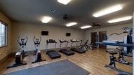 This single-story fitness room offers a clean, functional layout designed to support a variety of cardio-focused workouts. The space is outfitted with a row of treadmills as the centerpiece, complemented by an elliptical trainer and a stationary bike, providing users with multiple cardiovascular exercise options. Two flat-screen televisions are mounted on the walls, allowing exercisers to stay entertained during their workouts. The flooring consists of warm hardwood planks throughout the main exercise area, with interlocking rubber mat tiles placed near the entry zone, offering a dedicated area for stretching or floor exercises. Two ceiling fans are installed overhead, promoting air circulation and comfort during use. Recessed flush-mount light fixtures provide bright, even illumination throughout the room. The walls are finished in a neutral tone, contributing to a clean and focused atmosphere. Near the far end of the room, a set of wooden doors leads to an adjacent area, suggesting additional space beyond the fitness room itself. Overall, the room presents a practical and organized environment well-suited for regular physical activity.