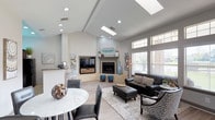 This single-story space showcases an open-concept layout that seamlessly connects the living and dining areas, creating a bright and inviting atmosphere. The vaulted ceiling rises dramatically overhead, enhanced by two skylights that flood the interior with natural light, complementing the recessed lighting throughout. Large windows along one wall offer views of the surrounding outdoors, further brightening the space.

The living area is anchored by a corner fireplace with a raised stone hearth and a coordinating mantel, providing both a functional and visual focal point. A flat-screen television is mounted nearby, making the space ideal for both relaxation and entertainment. Light hardwood-style flooring runs throughout, contributing to the airy, cohesive feel.

The dining area features a round marble-top table paired with sleek dark chairs, positioned adjacent to what appears to be a kitchen counter or bar area. The neutral gray-and-white color palette is accented with coastal-inspired blue décor elements, lending the space a contemporary yet welcoming character. Overall, the layout balances open flow with defined functional zones.