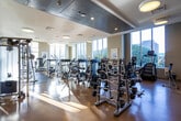 Located at 225 S Grand Ave in the heart of downtown Los Angeles, this modern fitness center offers a comprehensive and well-appointed workout facility. The space is thoughtfully organized into distinct zones designed to support a wide range of exercise routines. Large floor-to-ceiling windows line the perimeter, flooding the facility with natural light and offering striking views of the surrounding Los Angeles skyline and cityscape.

The main cardio and strength area is generously proportioned, housing a full lineup of Life Fitness treadmills, ellipticals, and stationary bikes, alongside a robust selection of selectorized weight machines and free-weight stations. Barbell racks, dumbbells, and cable machines are neatly arranged across polished hardwood-style flooring, with pendant drum lighting overhead adding a clean, contemporary feel.

A separate, glass-enclosed studio area provides a dedicated space for group classes, stretching, or personal training sessions. The studio features mirrored walls, a ballet barre, medicine balls, and a mounted flat-screen display. Its open floor plan and frosted-glass partitions lend a sense of privacy while maintaining visual connection to the broader facility.

Near the entry, a wooden cubby storage unit, hydration station, and thoughtful "LA"-branded signage contribute to the facility's polished, locally inspired aesthetic.