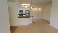 This single-floor residence showcases an open-concept layout that seamlessly connects the kitchen and living areas, creating a spacious and functional environment. The kitchen is well-appointed with stainless steel appliances, including a refrigerator, range, and microwave, all complemented by crisp white cabinetry and dark granite countertops. A curved breakfast bar serves as both a practical workspace and a natural divider between the kitchen and the living area, making it ideal for casual dining or entertaining. Pendant lights hang above the bar, adding a stylish focal point, while recessed track lighting along the ceiling provides additional illumination throughout the space. The flooring transitions from hardwood in the kitchen entry area to plush neutral carpet in the living space, offering a comfortable and inviting atmosphere. The light-toned walls and neutral color palette throughout contribute to a bright, airy feel. The open design of the layout allows for flexible furniture arrangements in the living area, maximizing the usable space and enhancing the overall sense of openness within the unit.