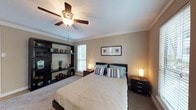 This bedroom is designed with both comfort and functionality in mind, presenting a warm, inviting atmosphere through its neutral beige color palette and soft carpeted flooring. Crown molding lines the upper perimeter of the room, adding a refined architectural detail that elevates the overall aesthetic. A ceiling fan with an integrated light fixture is centrally positioned, providing both air circulation and ambient lighting.

A standout feature of the room is the large, dark-toned built-in shelving unit along one wall, offering generous display and storage space. Decorative items and greenery are arranged across its shelves, contributing a personalized, lived-in feel. A window with horizontal blinds allows natural light to filter into the space, brightening the room during daytime hours.

The sleeping area is anchored by a dark-framed queen-sized bed dressed in neutral bedding accented with teal and striped pillows. Matching dark-wood nightstands flank the bed on both sides, each topped with warm-toned table lamps that cast a soft, welcoming glow. A framed piece of artwork above the bed adds a finishing decorative touch, completing the cohesive and well-balanced design of the room.
