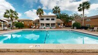Located at 2355 W Michigan Ave in Pensacola, Florida, this outdoor amenity area offers a well-appointed communal space designed for relaxation and recreation in a tropical setting. The centerpiece of the space is a generously sized swimming pool with crystal-clear blue water, marked with depth indicators and a lane divider, surrounded by a textured aggregate concrete deck that provides both durability and comfort underfoot. Lounge chairs are positioned throughout the pool deck, offering ample space for sunbathing and outdoor leisure. A white-painted covered pavilion with a gabled roof sits at one end of the deck, framed by tall swaying palm trees, mature oaks, and manicured shrubbery, creating a shaded retreat ideal for gathering. The surrounding residential buildings feature classic brick construction with balconies and large windows that overlook the pool area. A black iron perimeter fence encloses the space, providing both security and a clean visual boundary. The lush Florida landscape, combined with the open sky views and abundant natural light, gives the entire area a resort-like ambiance. This thoughtfully designed outdoor space reflects the warm, laid-back character of Pensacola living, making it a standout communal feature of the property.