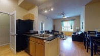 This thoughtfully designed 1-bedroom, 1-bathroom residence offers a functional and comfortable single-floor layout. The open-concept kitchen flows seamlessly into the living area, anchored by a central island with dark granite countertops, a double sink, and ample light wood cabinetry. Black appliances, including a range, microwave, refrigerator, and dishwasher, complete the kitchen. A dedicated laundry area is accessible just off the kitchen. The living room is warm and inviting, featuring hardwood-style flooring, a ceiling fan, and a large window dressed with teal curtains that allows plenty of natural light and outdoor views. The bedroom offers plush carpeting, a generously sized window with outdoor views, and space for both sleeping and a dedicated study or workspace. The bathroom is accessible from the bedroom and showcases warm tile flooring, an extended wood-toned vanity with ample cabinet storage, and a well-lit mirror. The entry foyer provides a clean transition into the open living space and is accented with track lighting above the kitchen island. Throughout the unit, warm neutral tones, elevated ceilings, and consistent finishes create a cohesive and comfortable living environment.