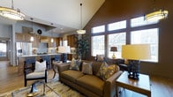 This single-floor residence showcases a thoughtfully designed open-concept layout that seamlessly connects the living area and kitchen into one cohesive, flowing space. The most striking architectural feature is the dramatic vaulted ceiling that rises to a steep peak, lending an impressive sense of height and grandeur to the main living area. A large bank of windows floods the space with abundant natural light, offering views of the surrounding neighborhood and creating a bright, welcoming atmosphere.

The living area is anchored by a generously sized sofa in warm earth tones, complemented by an array of decorative throw pillows in rich patterns of gold, brown, and blue. Layered lighting — including pendant fixtures, table lamps with warm amber shades, and recessed ceiling lights — creates an inviting ambiance throughout the day and evening.

The kitchen is visible from the living area and features warm wood cabinetry, a decorative tile backsplash, a custom range hood, and a peninsula with bar seating. Pendant lighting over the kitchen area ties the two spaces together cohesively. Light hardwood flooring runs throughout, grounding the rich, earthy color palette of chocolate browns, golds, and creams. The overall aesthetic is refined yet comfortable, blending contemporary design with traditional warmth.