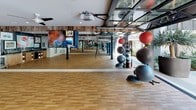 This single-level fitness studio is thoughtfully designed to accommodate a wide range of group exercise and training activities. The open floor plan is anchored by smooth hardwood flooring throughout, providing a durable and attractive surface suitable for dance, barre, yoga, and general fitness classes.

Full-length mirrors line the walls, enhancing the sense of space while allowing participants to monitor their form during workouts. Mounted barre rails run along the mirrored walls, making the studio well-suited for barre and dance-based instruction. Dark structural columns punctuate the open floor, adding an industrial contrast to the otherwise clean, modern aesthetic.

Reclaimed wood detailing along the ceiling adds warmth and texture, complementing the neutral white walls accented with slate-blue panels displaying framed artwork. A built-in counter area with wood cabinetry provides functional storage and workspace. Large glass panels and garage-style doors open toward a bright outdoor greenspace, flooding the studio with natural light and outdoor views.

Equipment storage is neatly integrated, with stability balls, resistance tools, and other accessories organized along the perimeter. Ceiling-mounted projectors and flat-screen TVs support multimedia-driven classes. Ceiling fans and exposed ductwork complete the studio's practical, well-ventilated design.