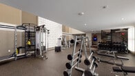 This two-floor fitness center is thoughtfully designed to accommodate a full spectrum of workout needs. The lower level serves as the primary strength training area, featuring rubber flooring throughout for safety and durability. A striking three-dimensional quilted white accent wall anchors the space visually, complemented by warm taupe wall finishes and recessed lighting overhead. This level houses an extensive array of strength equipment, including Life Fitness cable machines, a Synrgy360 functional trainer, multiple selectorized weight machines, and a well-stocked free weight section with a full dumbbell rack and adjustable benches. Large mirror panels along the walls enhance the sense of space and allow users to monitor their form. The upper level transitions to warm hardwood flooring and features bright, linear recessed lighting for a clean, modern aesthetic. This floor is dedicated to cardiovascular equipment, including a row of Life Fitness treadmills and an elliptical trainer. A curved black metal staircase connects the two floors with architectural elegance, while an elevator provides additional access. Together, both levels create a cohesive, well-appointed fitness environment suitable for a broad range of training styles.
