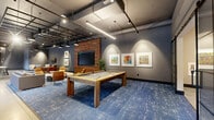 This generously sized single-floor amenity space blends industrial and contemporary design elements to create a dynamic, multi-functional environment. The open layout is anchored by polished concrete floors and high exposed ceilings with black steel grid framework, visible ductwork, and track lighting that add an urban, loft-inspired character throughout.

The main lounge area features rich blue patterned area rugs defining distinct seating zones, including plush upholstered armchairs, a round dining table with vibrant orange chairs, and a cozy TV nook framed by a full reclaimed wood accent wall with a large flat-screen display. A centerpiece pool table in natural wood adds a recreational focal point, while framed artwork lines the deep gray walls for a curated, gallery-like atmosphere.

A connected gathering room with exposed wood ceiling beams offers additional flexible seating with long communal tables and lounge chairs, ideal for meetings or social events. The fully equipped kitchen impresses with warm wood cabinetry, stainless steel appliances including a large refrigerator and double wall oven, a bold graphic tile backsplash, open shelving, and a substantial island with a stone countertop and bar seating. Oversized black-framed glass doors and windows along one wall flood the space with natural light and provide views to the surrounding courtyard.