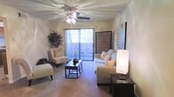 This single-floor space offers a welcoming and well-appointed interior designed for comfortable everyday living. The living room serves as the heart of the space, featuring plush wall-to-wall carpeting that adds warmth underfoot. A ceiling fan with integrated lighting anchors the room from above, providing both air circulation and ambient illumination. The room is furnished with a relaxed sofa accented with decorative pillows, a complementary accent chair with an ottoman, and a sleek dark coffee table that grounds the seating arrangement. Decorative touches, including framed artwork and a tall floor lamp, contribute to the room's inviting atmosphere. A standout feature is the full-height sliding glass door dressed with vertical blinds, which floods the space with natural light and provides direct access to a private outdoor balcony. The balcony, visible through the glass, offers a compact outdoor retreat with space for seating. Textured ceilings add a classic architectural detail throughout. Overall, the space strikes a balance between practicality and comfort, creating a bright and livable environment suited for relaxed daily life.