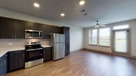 Located at 1700 W Frankford Rd in Carrollton, TX, this single-floor unit offers a thoughtfully designed open-concept layout that seamlessly connects the kitchen and living area. The kitchen is a standout feature, outfitted with rich dark espresso cabinetry, light quartz countertops, a white subway tile backsplash, and a full suite of stainless steel appliances including a refrigerator, electric range, over-the-range microwave, and dishwasher. Warm wood-look flooring flows throughout the main living areas, complementing the clean, neutral color palette.

The living space is bright and airy, benefiting from multiple windows dressed with horizontal blinds that frame pleasant outdoor views of surrounding greenery. A ceiling fan adds both function and style overhead. A glass-paneled entry door with sidelights allows additional natural light to fill the space.

The bedrooms are carpeted for comfort, each equipped with ceiling fans and windows overlooking the scenic surroundings. The bathroom features a full soaking tub and shower combination with white tile surround, a dark wood vanity with a white countertop, and wood-look flooring that ties the space together cohesively. The overall design is modern, clean, and functional.