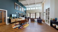 Located at 4401 Sprenkle Ln in Henrico, Virginia, this single-story community clubhouse delivers an inviting and well-appointed amenity space designed for both relaxation and activity. The centerpiece is a grand open lounge with soaring double-height ceilings accented by a coffered white ceiling structure and a bold teal feature wall adorned with large-scale framed artwork. Rich hardwood-style flooring runs throughout, anchoring vibrant area rugs that define distinct seating and social zones. A casual dining area with a round table and leather chairs sits alongside a lounge zone furnished with plush sofas and accent chairs. Built-in open shelving flanks a mounted flat-screen TV, creating a cozy media nook. A coffee and refreshment bar adds everyday convenience, while floor-to-ceiling windows and glass doors flood the space with natural light and offer views of the outdoor pool area. The fitness center is expansive and thoroughly equipped, featuring treadmills, ellipticals, stationary bikes, a cable machine, weight bench, dumbbell rack, and stability balls — all set against bold red accent walls, mirrored walls, and large windows that bring in natural light. Together, these spaces offer a dynamic and functional environment for community living.