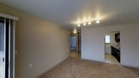 This single-floor residential space offers a clean, functional layout designed for comfortable everyday living. The main living area is generously proportioned, featuring wall-to-wall carpeting in a neutral tone that lends warmth to the interior. Smooth, light-colored walls and a white ceiling contribute to a bright and open atmosphere throughout the space.

Track lighting is a consistent design element, providing adjustable, focused illumination across the living areas. A sliding glass door fitted with vertical blinds allows natural light to filter in while offering flexible privacy control. An additional window in the main area further enhances natural light and ventilation, accompanied by a wall-mounted climate control unit for year-round comfort.

The layout transitions seamlessly from the main living area into a hallway that connects to additional rooms, suggesting a practical and well-organized floor plan. A ceiling-mounted flush light in the hallway ensures the connecting spaces remain well-lit. The continuity of carpeting and neutral wall tones throughout creates a cohesive and inviting aesthetic. Overall, this space is characterized by its simplicity, functionality, and readiness to be personalized into a comfortable home.