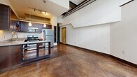 Located at 101 E Sixth St in the heart of Winston-Salem, NC, Unit 103 offers an industrial-modern aesthetic that makes a strong impression from the moment you enter. The open-concept layout seamlessly connects the kitchen and living area, creating a versatile and efficient use of space throughout the single-floor unit.

The kitchen is well-appointed with rich dark wood cabinetry, stainless steel appliances including a refrigerator and range, granite countertops, a center island with additional storage, and pendant lighting — all complemented by track lighting overhead. The living area benefits from soaring ceilings and large, multi-pane industrial-style windows that invite abundant natural light while offering a nod to the building's architectural heritage. Exposed ductwork and structural beams add to the loft-like character, while a ceiling fan provides comfort year-round.

Polished and stained concrete floors run throughout, lending a cohesive, urban feel to every room. The sleeping area is served by dual closets with bifold doors and wire shelving systems, offering practical and well-organized storage. A hallway connects the living spaces with ease.

The unit's industrial design details, high ceilings, and thoughtful finishes make it a distinctive urban residence in downtown Winston-Salem.