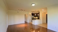 This single-floor space showcases an open-concept layout that seamlessly connects the living area to the kitchen. The living area features warm hardwood flooring that extends throughout, complemented by track lighting that provides adjustable illumination across the space. Neutral, light-toned walls contribute to a bright and airy atmosphere.

The kitchen is a standout feature, outfitted with rich dark wood cabinetry, stainless steel appliances including a refrigerator and range, and a granite-topped peninsula that doubles as a breakfast bar. Pendant lights hang above the peninsula, adding a refined touch to the cooking and dining area. A built-in sink is integrated into the peninsula, enhancing its functionality. Additional track lighting above the kitchen ensures the workspace is well-lit.

An arched architectural detail frames the transition between spaces, adding a subtle design element to the otherwise clean, modern interior. A hallway visible in the background leads to additional areas of the unit, hinting at further rooms beyond the main living and kitchen space. Overall, the layout feels spacious, functional, and well-designed for everyday living.