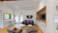 Located at 8401 Skillman St in Dallas, TX, this thoughtfully designed single-story space combines modern aesthetics with warm, natural materials to create an inviting and functional environment. The open-concept layout is anchored by soaring vaulted ceilings with exposed wood beam detailing, lending an airy, spacious feel throughout.

The main lounge area is a standout feature, showcasing a striking built-in media wall crafted from warm wood with a herringbone pattern backdrop, integrated shelving, and sleek white cabinetry below. A dark leather sofa, modern accent chairs, and layered textile wall art contribute to the space's curated, contemporary character. Light hardwood flooring flows seamlessly throughout, complementing the clean white walls.

A built-in counter with bar seating lines one wall of the lounge, framed by wood-trimmed windows that draw in abundant natural light and offer views of the surrounding outdoor area. A glass-paned door provides direct access to an outdoor patio space.

Adjacent to the main lounge is a compact kitchenette area featuring white countertops, a stainless steel sink, mosaic tile backsplash, and a built-in wood bench dining nook with a white table. Wood-framed windows flood this area with natural light and greenery views, creating a bright and welcoming atmosphere ideal for casual gatherings.