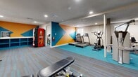 Located at 3304 Golden Rd in Tyler, TX, this fitness center is a well-equipped, single-level facility divided into two dedicated exercise rooms. The space is designed with functionality and energy in mind, featuring a vibrant color scheme highlighted by bold teal, orange, and gold geometric accent walls that create a motivating atmosphere throughout.

The main exercise room is outfitted with a variety of cardio equipment, including multiple treadmills, elliptical trainers, a rowing machine, and upright and cycling bikes. Full-length mirrors line portions of the walls, enhancing the sense of openness and allowing users to monitor their form. Durable teal rubber flooring covers the active zones, while patterned carpet tiles define the weight training area, where a rack of dumbbells, an adjustable weight bench, and a medicine ball storage rack provide a complete strength training setup.

The second exercise room offers additional cardio-focused equipment, including stationary bikes and low-impact machines, finished with the same teal rubber flooring. A water fountain is conveniently mounted on the wall nearby. Recessed lighting throughout both rooms provides consistent, bright illumination, and a textured ceiling adds a classic finishing touch. A Coca-Cola vending machine is available for refreshment. The overall layout is practical, clean, and purpose-built for a comprehensive workout experience.