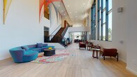 Located at 2020 Morton St in Charlotte, NC, this thoughtfully designed space makes an immediate impression with its grand, double-height lobby. Floor-to-ceiling windows and glass curtain walls flood the entry with natural light, while a striking textured brick accent wall and oversized glass doors establish a welcoming architectural identity. A sweeping staircase with a sleek metal railing anchors the central atrium, above which dramatic hanging sculptural artwork in vivid reds, oranges, and blues adds an artistic focal point.

The main lounge area features a curved teal sectional sofa positioned atop a vibrant, multicolored area rug, complemented by an ornate round coffee table. Adjacent seating nooks provide more intimate settings, including a retro-inspired arrangement of cushioned swivel chairs beneath a bold dome pendant light, set against a natural stone accent wall with outdoor views. A meditative lounge zone features low-profile dark sofas arranged around rustic reclaimed wood block tables, separated from neighboring areas by elegant floor-to-ceiling vertical wood slat partitions.

Throughout, light hardwood flooring, recessed lighting, curated wall art, and built-in wood benches contribute to a cohesive, contemporary aesthetic that balances vibrancy with comfort.