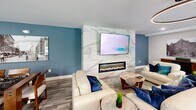 Located at 303 Dunbar St in Manchester, New Hampshire, this thoughtfully designed community lounge space occupies a single open floor. The layout seamlessly integrates multiple functional zones, creating a versatile environment suited for both relaxed gatherings and productive work sessions.

At the heart of the space is a striking kitchen area anchored by a large waterfall-edge quartz island with an undermount sink and pendant lighting overhead. White shaker-style cabinetry with matte black hardware lines the walls, complemented by a marble-look tile backsplash, a stainless steel side-by-side refrigerator, and a built-in microwave. A refrigerated snack and beverage station branded "The Edge" adds a convenient self-serve amenity near the dining area.

The lounge section features a bold accent wall clad in large-format marble-look tile, housing a sleek linear electric fireplace beneath a wall-mounted television — creating a refined focal point flanked by cream sofas and upholstered seating in rich navy blue tones. Adjacent to this is a casual work and dining area with velvet green booth-style seating, white pedestal tables, and vintage-style photographic artwork depicting local Manchester streetscapes.

Light wood-look flooring runs throughout, tying the zones together cohesively. Natural light filters in through a street-facing window, while recessed and ceiling-mounted fixtures provide consistent illumination across the entire space.