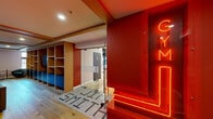 Located at 375 Rue de la Concorde in Montréal, the Le Smith amenity level offers residents a thoughtfully designed space combining fitness and relaxation. The entrance makes an immediate impression with a bold red accent wall, glowing neon "GYM" signage, a mosaic tile floor bearing the building's name, and built-in wood shelving with a blue seating nook — setting an energetic and stylish tone throughout.

The expansive exercise room is a standout feature, boasting soaring ceilings with an exposed dark ceiling structure and sleek suspended linear lighting. Light hardwood floors run throughout, complemented by floor-to-ceiling mirrors that amplify the sense of space. Large windows invite natural light and offer street-level views of Montréal. The fitness area is comprehensively equipped with treadmills, ellipticals, stationary bikes, cable machines, benches, free weights, and specialty training equipment, catering to a wide range of workout preferences.

Adjacent to the gym is a comfortable lounge area, finished with the same warm hardwood flooring and wood beam ceiling details. Deep tufted leather sofas and accent chairs surround a dark coffee table, creating an inviting space to unwind. A built-in shelving unit with bold red-painted interiors and a centrally mounted television serves as the room's focal point, blending function with distinctive design character.