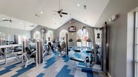 This fitness center is a thoughtfully designed, single-floor facility that combines architectural charm with a comprehensive array of exercise equipment. The space is defined by its striking vaulted ceilings, which lend an open, airy feel and are complemented by multiple ceiling fans that promote comfortable air circulation during workouts. Tall, arched windows along the walls flood the interior with abundant natural light while offering pleasant outdoor views of surrounding greenery and buildings, creating an inviting atmosphere for exercise.

The flooring features a bold, geometric patterned carpet in shades of blue and grey, adding a modern, energetic aesthetic to the space. Full-length mirrored walls on one side visually expand the room and provide functional feedback for form-conscious exercisers.

The cardio zone is well-appointed with multiple treadmills, elliptical trainers, and stationary bikes, all arranged to take advantage of the natural light and window views. The strength training area includes cable machines, a weight bench, and a freestanding dumbbell rack, catering to a variety of resistance training needs. Wall-mounted televisions are positioned throughout for entertainment during workouts.

Recessed lighting supplements the natural light, ensuring the space remains bright at all hours. The overall ambiance is clean, spacious, and motivating, making this fitness center a well-rounded destination for residents seeking a full workout experience.