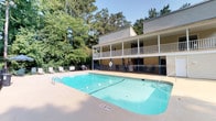 This outdoor recreational space centers around a generously sized swimming pool with clear, blue-green water, framed by a wide concrete deck that provides ample room for lounging and circulation. The pool is equipped with safety features including a life ring station, and the deck accommodates outdoor seating and umbrella-shaded areas for relaxation. A wrought-iron style fence lines the perimeter of the pool area, offering both safety and a clean visual boundary. The surrounding landscape is defined by tall, mature trees that provide natural shade and privacy, creating a serene, park-like atmosphere. Adjacent to the pool, a multi-level building features a covered upper balcony with railing, offering an elevated vantage point overlooking the pool and greenery below. The balcony's architectural design, with its traditional railing and column supports, complements the casual, resort-like character of the space. The building's light-toned exterior siding harmonizes with the natural environment. Overall, the space strikes a balance between functional amenity and relaxing outdoor retreat, well-suited for leisure and recreation in a nature-rich setting.