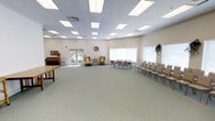 This single-floor space offers a generously sized, open multipurpose room designed to accommodate a variety of events and gatherings. The room features a neutral color palette of soft whites and warm beiges, creating a clean and welcoming atmosphere throughout.

The drop ceiling is fitted with an extensive grid of recessed fluorescent panel lights, ensuring bright, even illumination across the entire space. Large windows along one wall allow natural light to filter in, enhancing the room's airy, open feel. A glass-paned door provides additional natural light and outdoor access.

The room is carpeted in light, muted tones that complement the overall aesthetic. Seating is arranged flexibly, with rows of folding chairs set up on one side of the room, suitable for presentations or ceremonies. A pair of cushioned armchairs near the windows offer a more relaxed seating option. A upright piano and decorative framed artwork are visible along one wall, adding a touch of warmth and character.

Decorative wreaths and floral accents are placed throughout, contributing to the room's understated, tasteful ambiance. The open floor plan allows for easy reconfiguration to suit a wide range of uses.