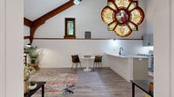 This remarkable two-floor space is a thoughtfully converted historic church that masterfully blends original architectural character with contemporary living. Spanning two levels, the lower floor houses the main kitchen and living areas, while the upper floor offers additional living space, a second kitchen, and a dedicated laundry room — all connected by two staircases.

The most striking features are the original stained glass windows, including a stunning floral rose window depicting a religious figure in vivid jewel tones, and a smaller arched stained glass window framed in rich dark wood trim. These windows cast colorful light throughout the interior, creating an ever-changing ambiance. Exposed dark wood beams trace the roofline and ceilings, preserving the sacred geometry of the original structure.

The modern kitchen is outfitted with gray shaker-style cabinetry, stainless steel appliances, white quartz countertops, and a matte black faucet — clean and functional against the historic backdrop. Light wood-look flooring flows throughout both levels, complementing the white walls and trim. The open living areas feel airy and generous beneath the soaring cathedral ceilings. A dedicated laundry room with a full-size washer completes the practical amenities. This is a truly one-of-a-kind space where history and modern comfort coexist seamlessly.