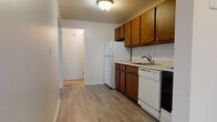 This single-floor unit offers a practical and functional layout with a clear distinction between living, sleeping, and utility spaces. The open living area is spacious and filled with natural light from a large window, featuring light gray vinyl plank flooring and neutral white walls. A textured ceiling and baseboard heating with a wall-mounted AC unit are present in the main living space, providing year-round climate control.

The kitchen is well-equipped with warm-toned wood cabinetry extending to the upper and lower sections, providing ample storage. It includes a full suite of white appliances — a refrigerator, dishwasher, and electric range — alongside a single-basin sink and light countertops. The kitchen connects seamlessly to the living area via an open pass-through column design.

The bedrooms are carpeted for comfort, each featuring windows that bring in natural light and baseboard heating. One bedroom includes a sliding-door closet with built-in shelving, offering practical storage solutions.

The bathroom is clean and functional, featuring fully tiled walls surrounding a soaking bathtub, a mosaic tile floor, a toilet, and a vanity with under-cabinet storage. A medicine cabinet above the vanity adds additional storage convenience. The overall ambiance is clean, bright, and move-in ready.
