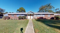 Located at 502 Ryan Ct in Sanford, North Carolina, this single-story brick building offers a classic and sturdy architectural presence. The exterior showcases traditional red brick construction accented by blue-gray shingle siding details beneath a gabled roofline, lending the structure a timeless, well-built character. A concrete walkway leads from the street directly to the front entrance, framed by a neatly maintained lawn and established shrubbery along the foundation beds.

The front entry features a white paneled door with a transom window above, a covered porch area, and landscaped mulched beds with young plantings that add curb appeal throughout the seasons. The building is part of the broader Sanford Estates community, as indicated by the neighborhood signage visible from the front, with similar brick structures lining the surrounding streets and mature trees providing a natural canopy backdrop.

The grounds are well-kept, with green lawns extending along the sides of the building and additional window units providing climate control. The porch area offers a sheltered outdoor space that opens to open views of the surrounding neighborhood. Overall, this property combines functional single-floor design with classic brick craftsmanship, situated within an established and cohesive residential community in Sanford, NC.