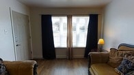 This single-floor space offers a comfortable and practical layout designed for everyday living. The living area is a standout feature, showcasing warm hardwood-style flooring that runs throughout and lends a cohesive, grounded feel to the room. Large windows dressed with horizontal blinds and layered curtains — combining sheer natural linen panels with bold dark drapes — allow for flexible light control while framing outdoor views of the surrounding neighborhood. The layered window treatments add both texture and depth to the space. Classic accent furniture, including ornately framed seating with neutral upholstery, contributes a traditional character to the room. A warm-toned table lamp provides soft ambient lighting, complementing the natural light that filters through the windows. Clean white walls and white baseboards keep the space feeling fresh and open, while the overall design strikes a balance between classic style and functional comfort. The single-floor layout offers ease of accessibility and a straightforward flow throughout. This space is well-suited for those seeking a cozy, traditionally styled environment with practical features and an abundance of natural light.