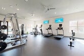 Located at 1633 Greenridge Drive in Burleson, TX, this single-floor fitness center offers a generously sized, open workout environment designed to accommodate a full range of exercise disciplines. The space features durable rubber flooring throughout, crisp white walls, and recessed lighting complemented by ceiling fans that maintain a comfortable atmosphere during workouts.

The cardio zone is equipped with multiple treadmills and elliptical machines, each fitted with individual display screens, and wall-mounted flat-screen TVs provide additional entertainment. A dedicated strength training area houses a multi-station cable machine, a smith machine with bench press setup, and a full rack of dumbbells arranged along a mirrored wall, allowing users to monitor their form with ease.

A separate functional training area features a freestanding punching bag, a wall-mounted display, and a dumbbell rack, bathed in natural light from arched windows that frame views of the surrounding greenery. An adjacent room provides additional space and connects to a restroom facility. Large glass doors open directly to the outdoor area, enhancing the sense of openness and providing convenient access to exterior amenities. The overall design is clean, modern, and highly functional, making this fitness center a comprehensive resource for residents.