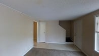 This two-floor residence offers a practical and straightforward layout designed for comfortable everyday living. The main level features wall-to-wall carpeting in the living area, which provides a warm and soft underfoot feel. Neutral beige walls and white trim throughout create a clean, cohesive aesthetic that lends itself well to a variety of décor styles. A textured popcorn ceiling adds a classic residential character to the space. The central staircase, framed by a clean drywall enclosure, serves as the primary connection between the two floors and acts as a natural focal point within the main living area. Multiple doorways are visible from the main room, suggesting a well-divided floor plan with distinct spaces for different functions. The transition between carpeted and hard-surface flooring near the entryway areas provides both practicality and visual separation between zones. Electrical outlets are positioned conveniently along the lower walls throughout the space. The overall ambiance is understated and move-in ready, offering a neutral canvas that can be easily personalized. The two-level configuration provides a natural separation between living and private spaces, making the home functional for a range of occupants.