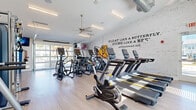 This fitness center is designed to inspire and accommodate a comprehensive workout experience. The space is anchored by a striking whitewashed brick accent wall bearing the iconic Muhammad Ali quote, "Float like a butterfly, sting like a bee," setting an energizing tone throughout. The open, single-level layout is generously proportioned, with soaring ceilings that lend an airy, expansive feel. Light wood-style flooring runs throughout, complementing the clean white walls and bright, modern aesthetic. Overhead, a combination of linear LED lighting and ceiling fans ensures optimal illumination and air circulation.

The cardio area is well-stocked with Matrix-brand treadmills, ellipticals, stair climbers, and stationary bikes, all arranged to maximize floor space. A multi-functional cable and strength training machine occupies one corner, accompanied by a free weight rack, an adjustable bench, and a punching bag. Resistance training equipment, exercise sliders, and stability balls round out the full-body fitness options. Large windows and a glass garage-style door flood the space with natural light and offer outdoor views, while a wall-mounted television adds entertainment. A connected anteroom with additional windows provides a transitional space within the facility.