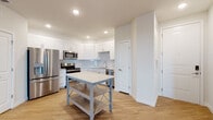 This well-appointed single-floor unit offers a clean, modern aesthetic throughout. The kitchen serves as a standout feature, showcasing crisp white shaker-style cabinetry, a classic subway tile backsplash, light stone countertops, and a full suite of stainless steel appliances including a French door refrigerator with ice maker, electric range, over-the-range microwave, and dishwasher. A freestanding island with a light countertop and lower shelf provides additional prep and storage space. Light hardwood-style flooring runs throughout the kitchen and open living area, which is bright and airy thanks to recessed lighting and a neutral white color palette. Double doors in the living area offer convenient closet or storage access. The bedrooms feature plush wall-to-wall carpeting, ceiling fans with light fixtures, and windows that allow natural light to filter in. At least one bedroom includes access to a walk-in closet. The bathroom is finished in a clean, minimalist style with a tub-shower combination, wood-look flooring, and white fixtures and vanity. Overall, the unit presents a fresh, move-in-ready interior with consistent finishes and a cohesive, contemporary design throughout.