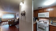This single-floor residence offers a practical and comfortable layout designed for everyday living. Rich, dark hardwood-style flooring runs consistently throughout the home, providing a warm and cohesive aesthetic from room to room.

The living room serves as the heart of the home, featuring a large window dressed with sheer curtains that invite natural light into the space. The room comfortably accommodates plush seating, a coffee table, and entertainment furnishings, creating a relaxed and inviting atmosphere. Wall-mounted décor and a framed mirror add character to the neutral-toned walls.

Adjacent to the living area, the dining space features a classic wooden table and chairs, positioned near the kitchen entrance for convenient flow. The kitchen is well-appointed with warm-toned wood cabinetry, granite-style countertops, a tile backsplash, and white appliances including a coil-top electric range, an over-the-range microwave, and a refrigerator.

The primary bedroom offers a calm retreat with a wooden sleigh bed frame and window treatments providing privacy. A second bedroom, featuring its own window with blinds, provides additional flexibility. The full bathroom includes a vanity with storage, a framed mirror, and a tub-shower combination, completing this functional and homey residence.