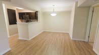 This single-floor space offers a clean, open layout designed for comfortable everyday living. The dining area is anchored by a sleek pendant light fixture that hangs from a smooth ceiling, casting a warm, focused glow over the space. Light-toned laminate wood flooring runs throughout, lending a bright and cohesive feel to the interior. Crisp white baseboards provide a polished contrast against the neutral, softly painted walls, contributing to an overall sense of cleanliness and simplicity.

A pass-through window connects the dining area to the kitchen, promoting an open flow between spaces. The kitchen features rich, dark espresso-toned cabinetry that adds depth and contrast to the lighter surroundings. A mosaic tile backsplash in earthy, varied tones serves as a visual focal point, adding texture and character. Stainless steel appliances, including what appears to be a microwave, complement the cabinetry and reinforce a modern aesthetic. A countertop bar ledge along the pass-through provides additional prep or casual seating space. Together, these elements create a functional, well-appointed interior with a cohesive and contemporary design sensibility.
