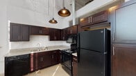 Located at 1700 Fairview St in Greensboro, North Carolina, this single-floor unit showcases a compelling industrial loft aesthetic rooted in its converted building origins. Soaring ceilings immediately establish a dramatic sense of vertical space, while painted exposed brick walls and polished concrete floors reinforce the raw, warehouse-inspired character throughout.

The kitchen is a standout feature, outfitted with rich dark-stained cabinetry, light stone-toned countertops, and a full suite of black appliances including a refrigerator, electric range, over-the-range microwave, and dishwasher. A double-basin sink adds functional depth, and industrial-style pendant lights hang overhead, complementing the space's urban character. A stainless steel work surface provides additional prep space. Large windows fitted with horizontal blinds allow natural light to pour in, offering views of the surrounding neighborhood.

The bathroom is clean and straightforwardly appointed, featuring a vanity with a white countertop, a toilet, and a soaker tub with a curtain rod and grab bar for accessibility. Neutral walls and polished concrete flooring maintain design consistency with the rest of the unit.

The open floor plan, as revealed in the dollhouse view, presents a well-organized layout with distinct kitchen, living, and bathroom zones, making this a versatile and characterful space.