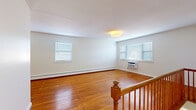 This single-floor unit showcases a clean, functional layout with warm hardwood flooring that runs consistently throughout the living and bedroom areas, lending a cohesive and inviting atmosphere to the space. The open living area is generously sized, benefiting from multiple windows fitted with white horizontal blinds that allow natural light to fill the room. A wall-mounted air conditioning unit and baseboard heating ensure year-round climate comfort.

The bedroom areas continue the hardwood flooring theme and are well-proportioned, each featuring baseboard heating and window access to natural light. Crisp white walls throughout maintain a bright, neutral palette suitable for a variety of interior styles. White panel doors with sleek black hardware add a modern contrast to the warm wood tones underfoot.

The kitchen is outfitted with white shaker-style cabinetry, light quartz countertops, a classic white subway tile backsplash, and stainless steel appliances including a gas range and over-the-range microwave — a practical and stylish cooking space.

The bathroom presents a clean, contemporary aesthetic with large-format light gray marble-look floor and wall tiles, a soaking tub with a tiled surround, and minimalist black fixtures, creating a calm and polished retreat. A staircase with a warm wood railing provides access to the floor.