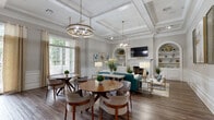 Located at 100 Ivey Park Lane in Norcross, Georgia, this beautifully appointed community clubhouse offers a refined gathering space defined by classic architectural details and upscale finishes. The single-floor layout flows seamlessly between distinct areas, all unified by rich hardwood-style flooring, soft neutral wall tones, and crisp white wainscoting that lends an air of timeless elegance throughout.

The main lounge is a standout, featuring soaring coffered ceilings with detailed crown molding and a pair of gold-toned geometric chandeliers that cast warm light across the space. A traditional white fireplace flanked by built-in arched shelving serves as the room's focal point, complemented by teal upholstered seating, gold-accented furnishings, and curated artwork. Adjacent to the lounge, a spacious dining area accommodates multiple round tables and modern upholstered chairs, ideal for casual gatherings.

A secondary lounge area opens to the outdoors through full-length French doors, flooding the space with natural light and offering views of the surrounding greenery. A bar-height counter with bar seating and a teal accent wall adds a vibrant, social atmosphere to this section. A connecting corridor with coffered ceilings links the spaces, with a refreshment station providing additional convenience. The overall ambiance is warm, sophisticated, and welcoming.