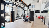This single-floor fitness center is designed to support a full spectrum of workout routines in a bright, open environment. The space is defined by its soaring ceilings and expansive floor-to-ceiling arched windows that line the perimeter walls, flooding the interior with abundant natural light and offering views of the surrounding exterior. Light hardwood flooring runs throughout, contributing to a clean and modern aesthetic.

The main training area is anchored by a large functional rig system equipped with suspension trainers, battle ropes, a heavy punching bag, and a plyo box, making it well-suited for high-intensity and functional fitness training. A dedicated strength zone features a cable and weight machine, a flat bench, and a well-stocked dumbbell rack. Cardio equipment, including multiple treadmills and stationary bikes, is arranged along the windowed walls to maximize natural light during workouts.

A striking large-scale mural of a dancer adorns one wall, adding an artistic focal point to the space. Floor-to-ceiling mirrors along one side visually expand the room and support form-focused training. A wall-mounted flat-screen TV, medicine balls, stability balls, and a weight plate storage rack round out the available amenities. Accessible restroom facilities and dual water fountains are conveniently located within the space.