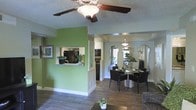 This single-story home showcases an open-concept design that effortlessly connects the living, dining, and kitchen areas into one cohesive space. The interior is characterized by bold green accent walls that add a vibrant, energetic feel, complemented by wood-style flooring that runs throughout, lending warmth and a sense of continuity.

The living area features a ceiling fan with a built-in light fixture mounted on a textured ceiling, providing both comfort and ambiance. A pass-through window connects the kitchen to the main living space, offering a convenient serving area while maintaining an open sightline. The kitchen, visible through the pass-through, is equipped with black appliances and ample cabinetry.

The dining area is thoughtfully appointed with a glass-top table surrounded by dark upholstered chairs, creating a sleek, modern aesthetic. A pendant light fixture hangs above, adding a refined focal point. Decorative mirrors lean against the dining room wall, amplifying natural light that streams in from a nearby window, enhancing the overall brightness and openness of the space.