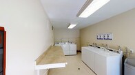 This single-floor communal laundry facility is designed with practicality and efficiency in mind. The room is finished with neutral beige walls and light-toned flooring, creating a clean and welcoming atmosphere. Overhead, bright fluorescent panel lighting ensures the entire space is well-illuminated throughout the day.

The facility is equipped with multiple top-loading washing machines and front-loading dryers arranged in rows along the walls, providing ample capacity for residents. A long, built-in folding counter runs along one wall, offering generous surface area for sorting and folding laundry with ease. Informational notices and posted guidelines are displayed on the wall near the machines, keeping users informed of proper usage and any facility rules.

The layout is thoughtfully organized, allowing multiple users to move around comfortably and access machines without congestion. The light color palette of the walls and floor contributes to a bright, hygienic feel that is easy to maintain. Overall, this laundry facility is a practical, no-frills space designed to serve the everyday needs of its users efficiently and comfortably.