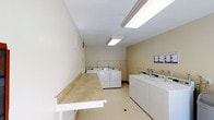 This single-floor communal laundry facility is designed with practicality and efficiency in mind. The room is finished with neutral beige walls and light-toned flooring, creating a clean and welcoming atmosphere. Overhead, bright fluorescent panel lighting ensures the entire space is well-illuminated throughout the day.

The facility is equipped with multiple top-loading washing machines and front-loading dryers arranged in rows along the walls, providing ample capacity for residents. A long, built-in folding counter runs along one wall, offering generous surface area for sorting and folding laundry with ease. Informational notices and posted guidelines are displayed on the wall near the machines, keeping users informed of proper usage and any facility rules.

The layout is thoughtfully organized, allowing multiple users to move around comfortably and access machines without congestion. The light color palette of the walls and floor contributes to a bright, hygienic feel that is easy to maintain. Overall, this laundry facility is a practical, no-frills space designed to serve the everyday needs of its users efficiently and comfortably.