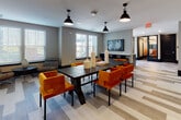 This thoughtfully designed single-floor amenity space offers a versatile and contemporary environment suited for both focused work and casual gathering. The main lounge area is anchored by a substantial dark wood communal table surrounded by plush, burnt-orange upholstered chairs, illuminated by industrial-style pendant lights that add a modern character to the space. Wide-plank, multi-toned luxury vinyl flooring runs throughout, lending a cohesive and polished aesthetic across all zones.

A dedicated lounge nook along the perimeter features large windows with horizontal blinds that invite abundant natural light, paired with eclectic accent seating and a wall-mounted display screen, creating a comfortable space for informal gatherings or individual work. A private conference room equipped with a round white table, high-back orange leather chairs, and textured accent wallpaper offers a refined setting for meetings, with natural light filtering through its generous windows.

A striking semi-private lounge section defined by bold navy blue accent walls and structural columns houses intimate booth-style seating areas with white oval tables and warm wood storage benches, accented by vibrant abstract artwork. The entry foyer features sleek dark double doors, light wood flooring, and a water station, completing this well-appointed and multi-functional space.