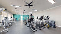This fitness center offers a thoughtfully organized workout environment spread across a single floor. The space is divided into distinct functional zones, each designed to support a different aspect of physical training. The cardio area features a lineup of True-brand ellipticals, stationary bikes, and treadmills, complemented by ceiling fans and recessed fluorescent lighting that keep the area comfortable during intense sessions. A wall-mounted television is positioned for easy viewing during workouts. The strength-training zone houses a robust selection of cable machines and selectorized equipment, arranged along a windowed wall that allows ample natural light to filter in through slatted blinds, offering views of the surrounding outdoor area. A dedicated free weights section includes a full dumbbell rack, a multi-functional rack with weight plates, and adjustable benches, set against a vibrant teal accent wall with mirrors to aid in form and technique. The flooring throughout consists of durable interlocking rubber mat tiles, providing traction and impact absorption. Crown molding adds a refined architectural touch to the space. French doors connect sections of the facility, and the overall layout ensures clear circulation paths between all equipment areas, creating a functional and inviting environment for users of all fitness levels.