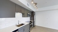 This 1-bedroom, 1-bathroom unit features a thoughtfully designed single-floor layout that maximizes functionality within a compact footprint. The kitchen serves as a standout space, showcasing a sleek, modern aesthetic with flat-front cabinetry in a warm charcoal-gray finish, complemented by clean white quartz countertops and a classic white subway tile backsplash. Stainless steel appliances, including a built-in microwave, electric range, and refrigerator, provide a polished, cohesive look. A stainless steel undermount sink with a contemporary faucet adds to the kitchen's refined details. Track lighting mounted on the ceiling offers targeted illumination, enhancing the modern atmosphere. Light-toned wood-style flooring runs throughout, lending warmth and continuity to the space. The overall design palette — combining neutral whites, cool grays, and natural wood tones — creates a clean, contemporary ambiance. The open layout between the kitchen and living area promotes an efficient flow, making the unit feel welcoming and functional for everyday living.